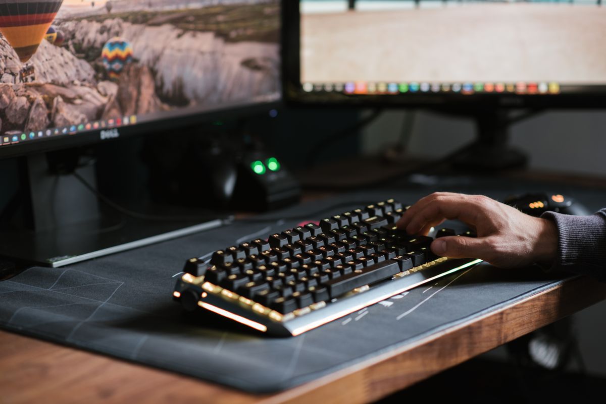 Close up view of a desk. A persons hand is visible typing on a keyboard. There are two monitors partially visible.